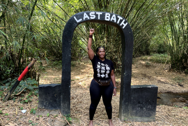 Woman standing in front of last bath inscription with right hand raised.